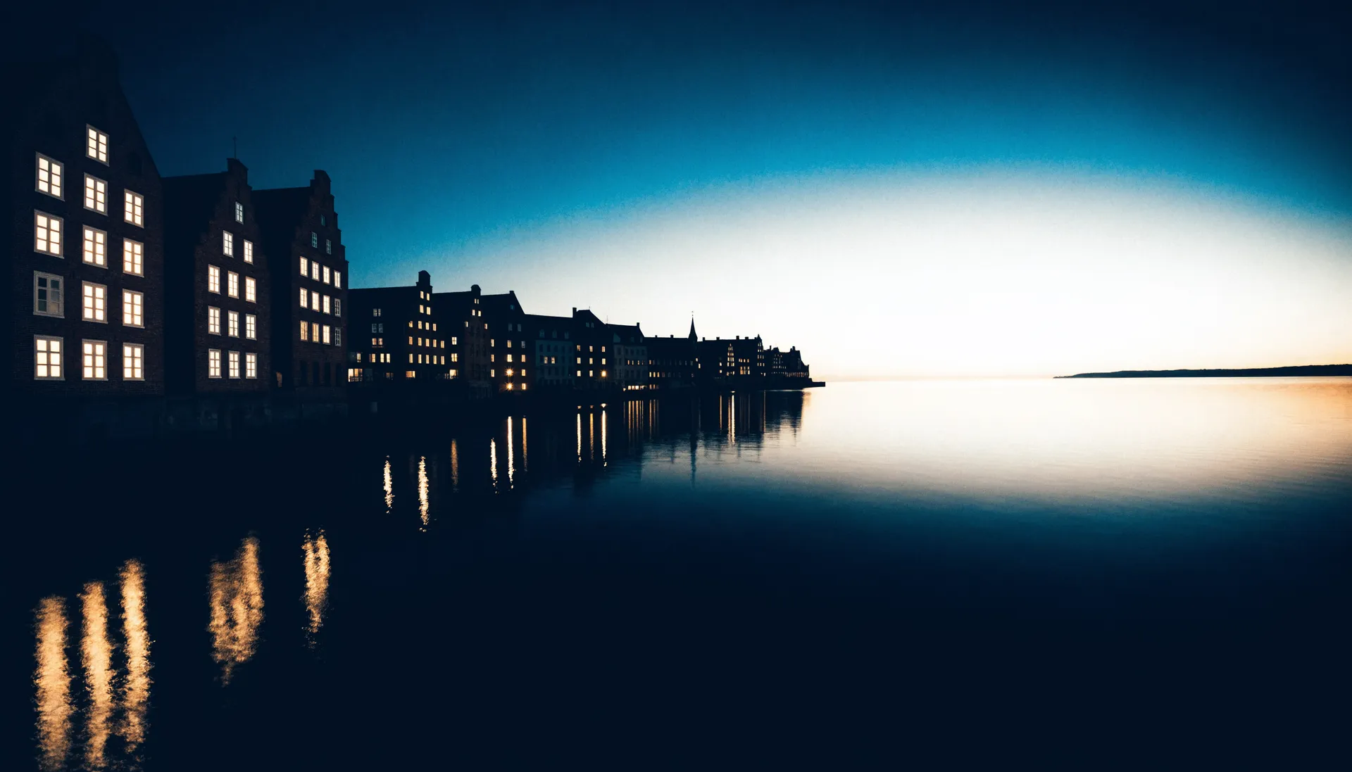 Hansestädtische Backsteinfassaden am Hafen zur blauen Stunde, Lichter spiegeln sich im Wasser.